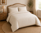 A neatly made bed in a warm, softly lit bedroom featuring a cream and beige vertically striped duvet cover and matching pillow shams. The bed has a light wood headboard and is styled with a wooden nightstand, a lamp, a framed lion portrait, and a potted plant.