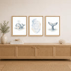 Three framed whale-themed wall art prints hang above a light wood console table in a styled room. The center print features a watercolor whale tail with the quote 'My plans are my plans.