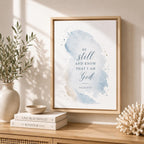 Framed wall art with a watercolor-style blue and beige wash and the text 'BE still AND KNOW THAT I AM God' on a light wood console table, styled with books, a bowl, a vase with greenery, and coral decor.