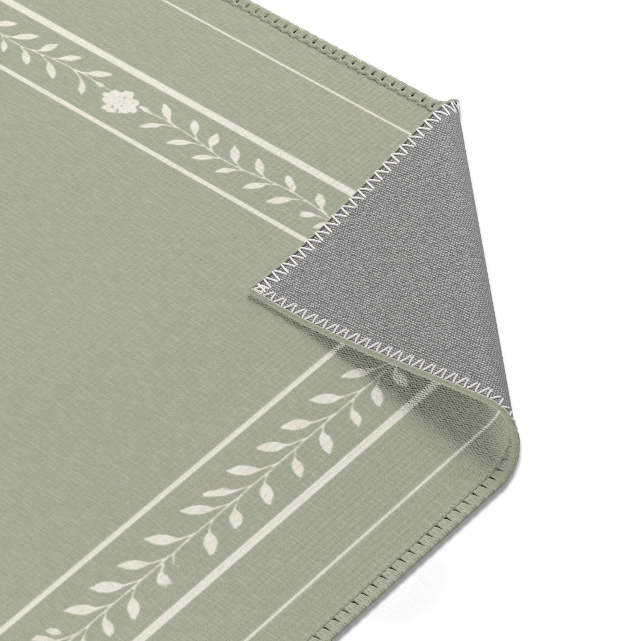 A close-up of a rectangular mat or rug with a sage green top, white leafy stripe pattern, and a gray underside showing stitched edges, photographed on a white background.
