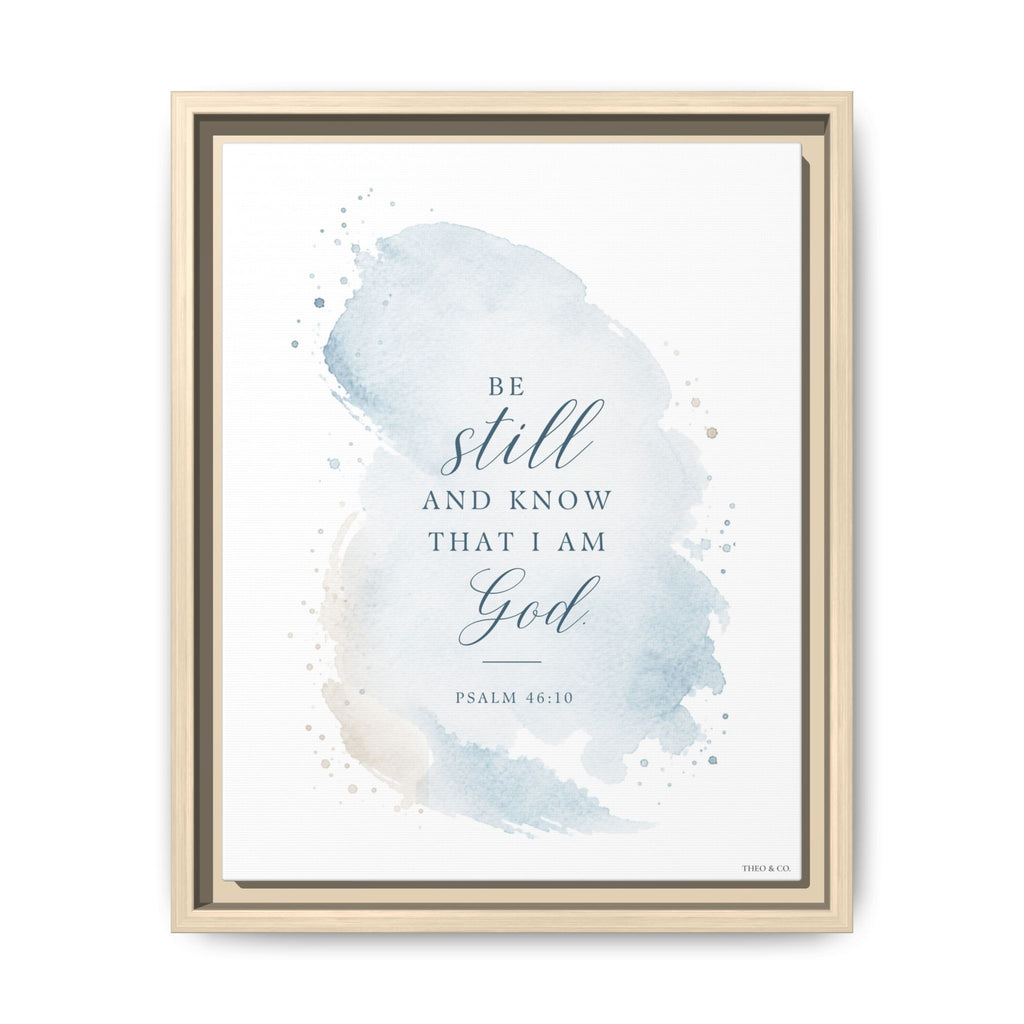 Framed wall art featuring a light blue watercolor splash with the Bible verse 'Be still and know that I am God' and 'Psalm 46:10' in dark blue text, shown in a light wood frame against a white background.