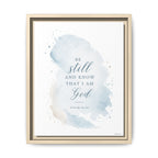 Framed wall art featuring a light blue watercolor splash with the Bible verse 'Be still and know that I am God' and 'Psalm 46:10' in dark blue text, shown in a light wood frame against a white background.