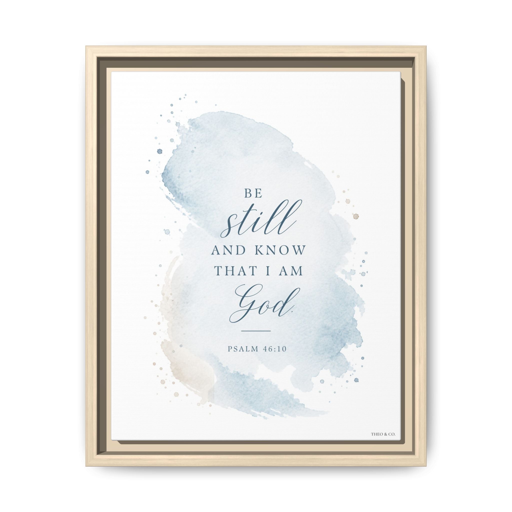 Framed wall art featuring a light blue watercolor splash with the Bible verse 'Be still and know that I am God' and 'Psalm 46:10' in dark blue text, shown in a light wood frame against a white background.