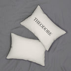 Two white rectangular throw pillows on a gray bedspread, with one pillow featuring the name 'THEODORE' printed in black serif lettering.