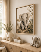 A framed wall art print showing a detailed illustration of a baby elephant, styled in a warm, neutral nursery or children's room above a wooden console table with a lamp, books, and a plush toy.