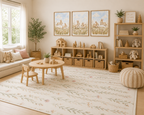A beautifully styled children's playroom featuring a large floral cream rug with a delicate green and pink floral vine pattern, a small round wooden table and chairs, wooden storage cubbies, a striped pouf, and three framed animal prints on the wall.