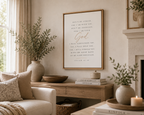 A framed vertical wall art print with a Bible verse in elegant black and gold script, styled in a warm, neutral-toned living room above a wooden console table.