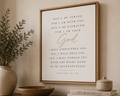 A framed wall art print featuring a Bible verse from Isaiah 41:10 in black serif text with 'God' in gold script, displayed in a light wood frame on a beige wall above a tabletop with a plant, bowl, and books.