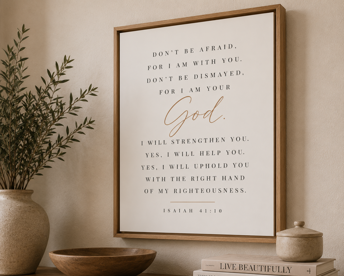 A framed wall art print featuring a Bible verse from Isaiah 41:10 in black serif text with 'God' in gold script, displayed in a light wood frame on a beige wall above a tabletop with a plant, bowl, and books.