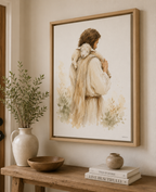 A framed art print depicting a man Jesus in a cream-colored cloak carrying a white lamb, styled on a wooden console table with a vase, bowl, and books in a warm, neutral-toned room.