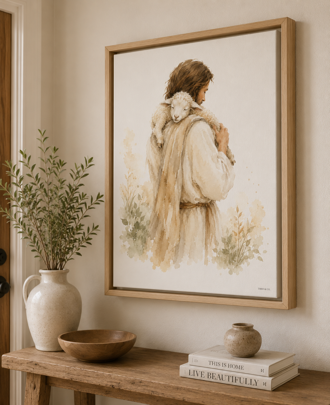 A framed art print depicting a man Jesus in a cream-colored cloak carrying a white lamb, styled on a wooden console table with a vase, bowl, and books in a warm, neutral-toned room.