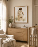 A framed art print depicting a man Jesus in a cream-colored cloak carrying a white lamb, styled on a wooden dresser table with a vase, bowl, and books in a warm, neutral-toned room.