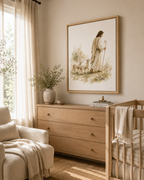 A framed art print depicting Jesus and sheep hangs on a beige wall in a softly lit nursery. The room includes a light wood dresser, a crib with a white blanket, a chair with a cream throw, and a window with sheer curtains.