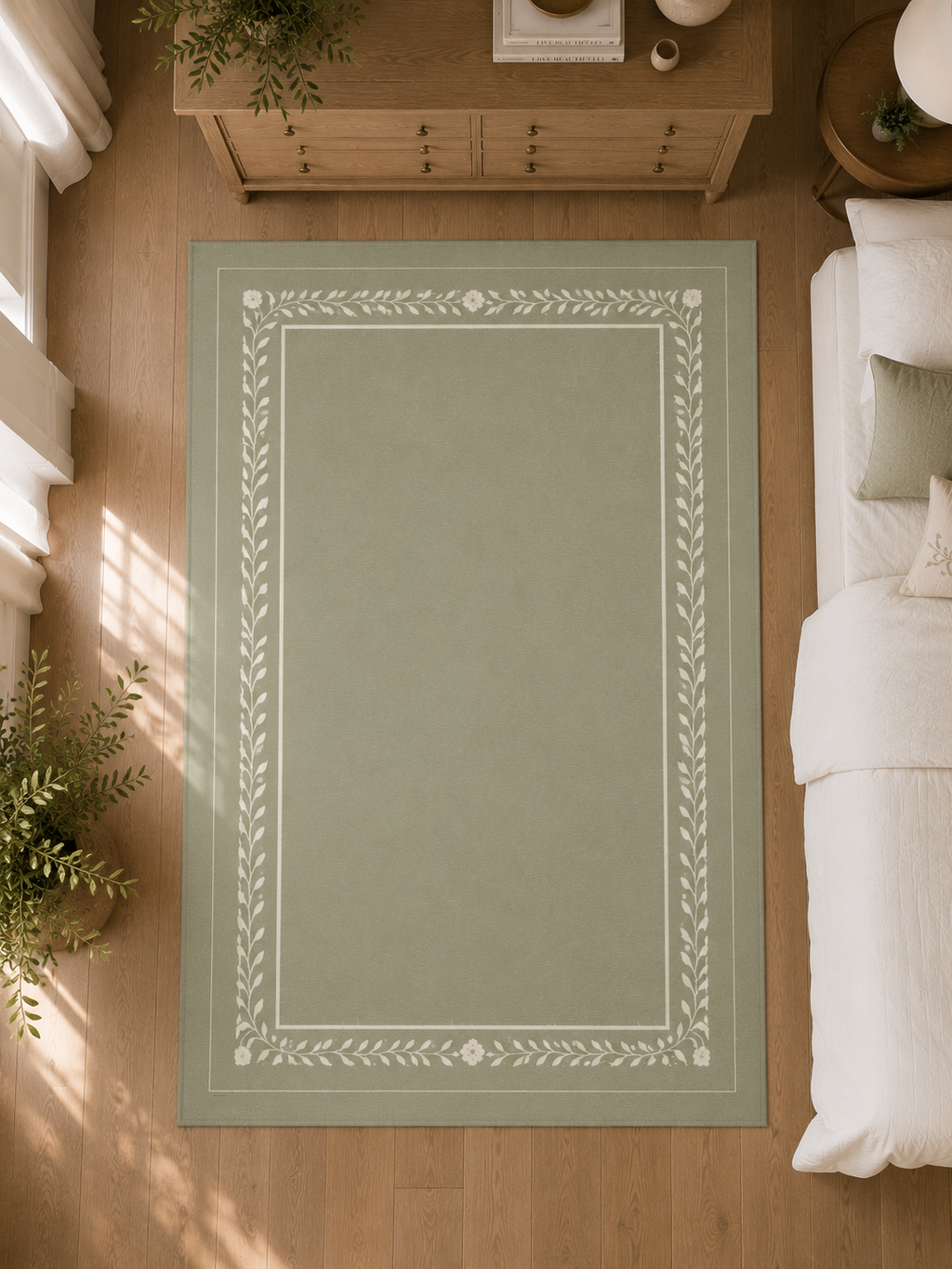 A rectangular area rug in a muted sage green color with a white decorative border, shown from a top-down perspective in a bedroom setting with a wooden floor, bed, and dresser.