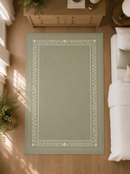 A rectangular area rug in a muted sage green color with a white decorative border, shown from a top-down perspective in a bedroom setting with a wooden floor, bed, and dresser.