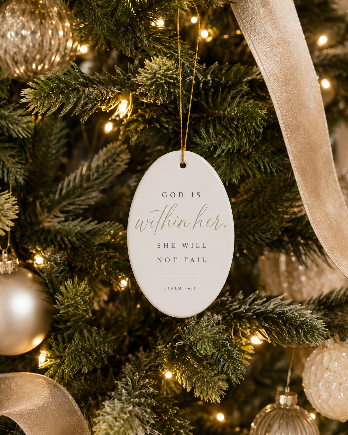 An oval white Christmas ornament with gold and black text reading 'GOD IS within her. SHE WILL NOT FAIL' and 'PSALM 45:3', hanging on a decorated Christmas tree with warm lights, silver ornaments, and a beige ribbon.