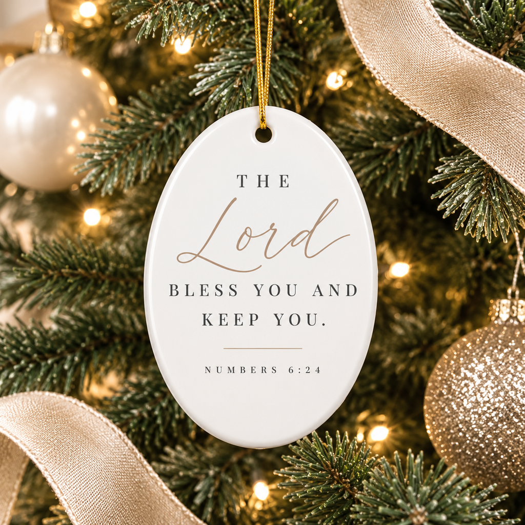 An oval white Christmas ornament hanging from a gold string on a decorated Christmas tree. The ornament features the printed text 'THE Lord BLESS YOU AND KEEP YOU.