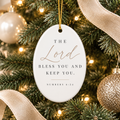 An oval white Christmas ornament hanging from a gold string on a decorated Christmas tree. The ornament features the printed text 'THE Lord BLESS YOU AND KEEP YOU.