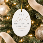 An oval white Christmas ornament hanging from a gold string on a decorated Christmas tree. The ornament features the printed text 'THE Lord BLESS YOU AND KEEP YOU.