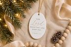 An oval white Christmas ornament with elegant gold and black typography reading 'The Lord Bless You and Keep You. Numbers 6:24', styled on a beige fabric background with pine branches, warm string lights, ribbon, wooden beads, and pinecones.