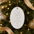 An oval white Christmas ornament with gold hanging string and printed scripture text, displayed on a decorated Christmas tree with green branches, warm lights, and gold ribbon.