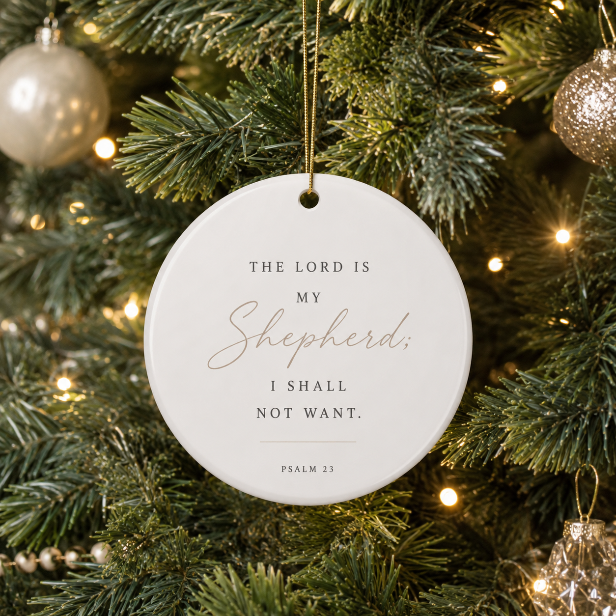 A round white ceramic Christmas ornament hanging from a gold string on a decorated Christmas tree. The ornament features the Bible verse 'THE LORD IS MY Shepherd; I SHALL NOT WANT.