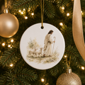A round white ceramic Christmas ornament featuring a painted illustration of a shepherd with sheep, hanging on a decorated Christmas tree with warm lights and gold ornaments.