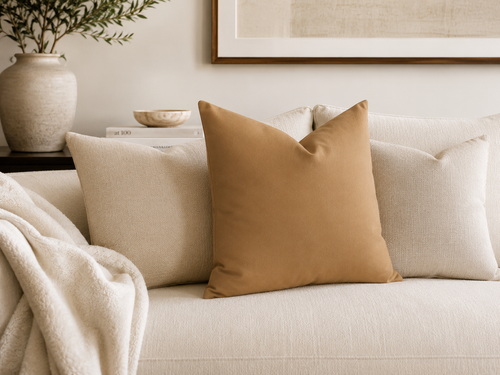 A tan throw pillow centered on a light beige sofa in a styled living room setting, with matching beige pillows, a cream throw blanket, a vase with greenery, and framed wall art in the background.