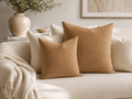 Two square tan throw pillows displayed on a cream-colored sofa in a styled living room setting with a neutral throw blanket, a ceramic vase, and framed wall art.