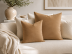 Two square tan throw pillows displayed on a cream-colored sofa in a styled living room setting with a neutral throw blanket, a ceramic vase, and framed wall art.