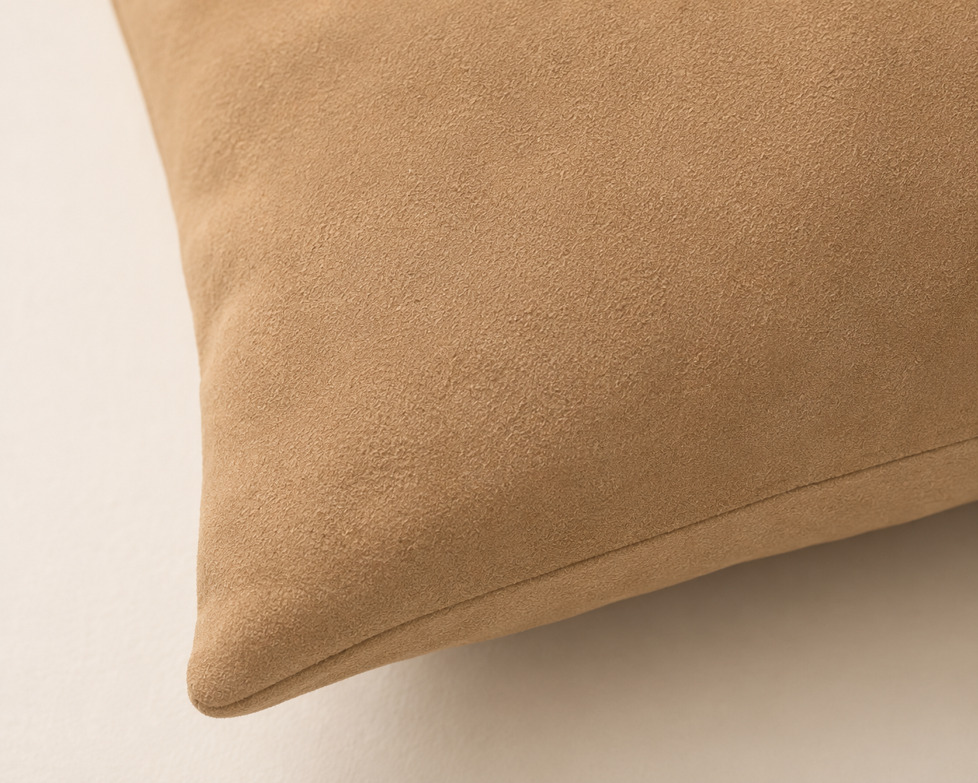 A close-up of a tan suede or microfiber throw pillow corner on a light beige background.