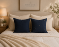 A styled bedroom scene with a bed made up in cream bedding and two navy throw pillows in front. One pillow is solid navy, and the other has a subtle gold star pattern.