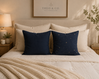 A styled bedroom scene with a bed made up in cream bedding and two navy throw pillows in front. One pillow is solid navy, and the other has a subtle gold star pattern.