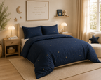 A navy blue bedding set with a starry night sky pattern is styled on a bed in a warm, cozy bedroom setting with wooden nightstands, lamps, and a beige rug.