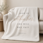 An off-white fleece throw blanket draped over a light beige sofa in a neutral living room setting. The blanket features the printed text 'GOD IS within her, SHE WILL NOT FAIL' and 'PSALM 46:5' in dark gray lettering, with 'within her' in a warm rose-gold script.