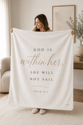 A woman holds up a large cream-colored fleece blanket in a bright, modern living room. The blanket features the Bible verse 'GOD IS within her, SHE WILL NOT FAIL' and 'PSALM 46:5' printed in black and rose gold lettering.
