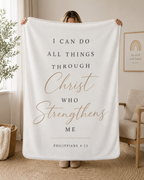 A person holds up a large white fleece blanket in a cozy, modern living room setting. The blanket features a printed Bible verse from Philippians 4:13 in black and rose gold lettering.