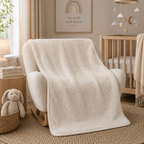 A plush cream-colored armchair in a styled nursery, draped with a matching textured cream throw blanket. The room features a wooden crib, a rabbit toy in a basket, and a framed 'be still and know' print on the wall.