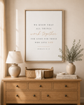 A framed typographic wall art print with a Bible verse hangs above a wooden dresser in a styled room. The print has a white background with black and blush pink text, a light wood frame, and is surrounded by home decor including a lamp, basket, books, and a vase with greenery.