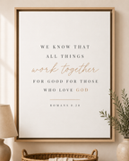 A framed inspirational wall art print featuring the Bible verse Romans 8:28, 'WE KNOW THAT ALL THINGS work together FOR GOOD FOR THOSE WHO LOVE GOD,' styled in a warm home setting with a lamp, basket, and plant.