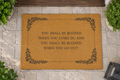 A natural coir doormat with a black printed decorative border and the phrase 'YOU SHALL BE BLESSED WHEN YOU COME IN, AND YOU SHALL BE BLESSED WHEN YOU GO OUT' placed on a stone walkway near plants.