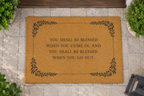 A natural coir doormat with a black printed decorative border and the phrase 'YOU SHALL BE BLESSED WHEN YOU COME IN, AND YOU SHALL BE BLESSED WHEN YOU GO OUT' placed on a stone walkway near plants.