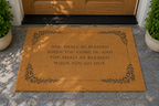 A natural coir doormat with black printed text and decorative leaf borders, placed in front of a wooden door on a stone walkway with potted plants on both sides.