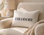A square off-white throw pillow with the name 'THEODORE' printed in black serif letters, resting on a textured cream armchair in a warm, sunlit room.