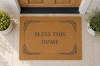 A natural coir doormat with the words 'BLESS THIS HOME' and a decorative leaf border, placed in front of a wooden front door on a tiled entryway floor, with potted plants on both sides.