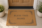 A rectangular coir doormat with the words 'BLESS THIS HOME' and a leafy border, placed in front of a wooden front door on a tiled entryway floor, with potted plants on both sides.
