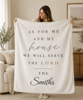 A woman holds up a large cream-colored fleece blanket in a cozy living room setting. The blanket features personalized printed text in black and muted gold, reading 'AS FOR ME AND MY house WE WILL SERVE THE LORD THE Smiths'.