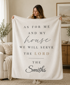 A woman holds up a large cream-colored fleece blanket in a cozy living room setting. The blanket features personalized printed text in black and muted gold, reading 'AS FOR ME AND MY house WE WILL SERVE THE LORD THE Smiths'.