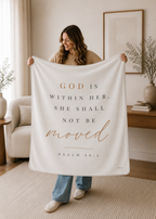 A woman in a beige sweater and blue jeans holds up a large cream throw blanket with the printed text 'GOD IS WITHIN HER, SHE SHALL NOT BE moved PSALM 46:5' in black and rose gold lettering, set in a bright, modern living room.