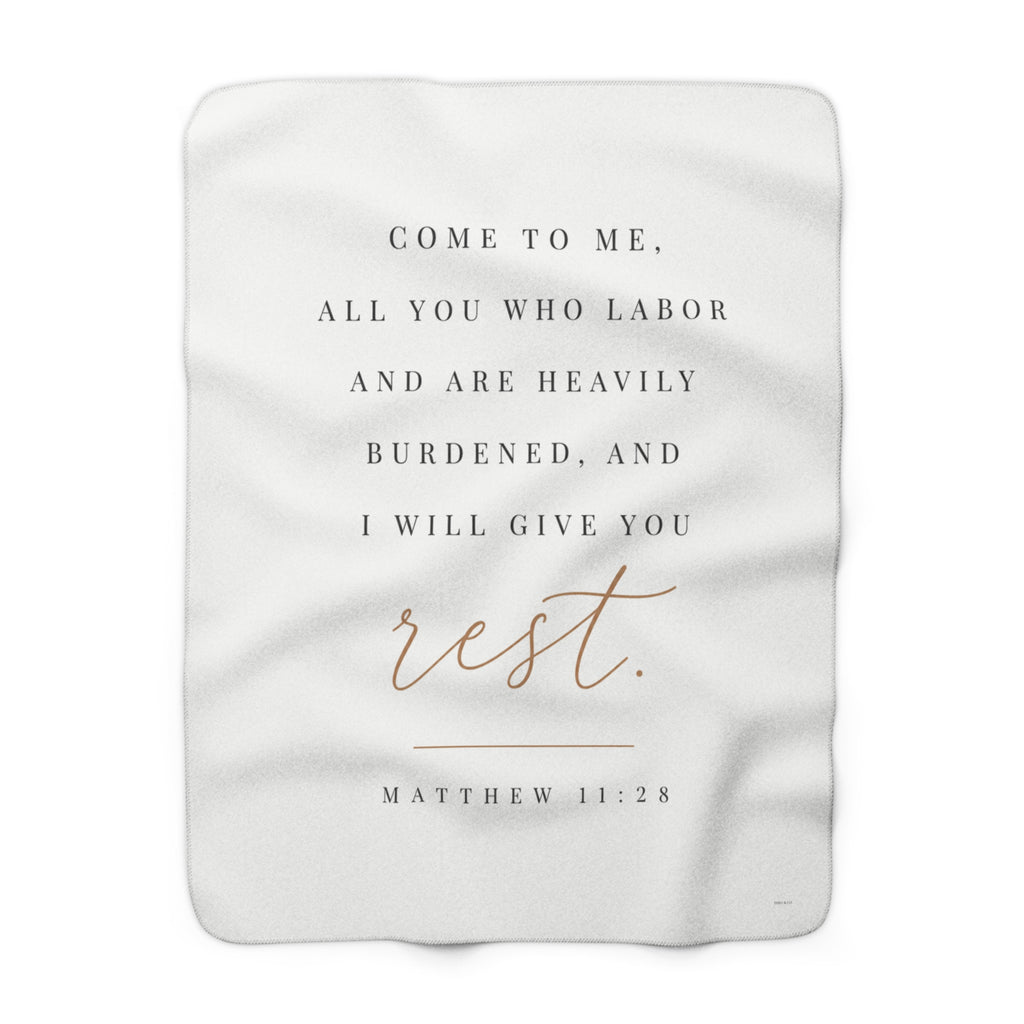 A white rectangular blanket with rounded corners featuring a printed Bible verse from Matthew 11:28. The text reads 'COME TO ME, ALL YOU WHO LABOR AND ARE HEAVILY BURDENED, AND I WILL GIVE YOU rest.